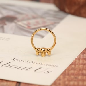 May include: A gold-colored nose ring featuring three small skull charms. The ring is circular and has a hinged closure. The skulls are detailed with eye sockets, noses, and teeth. The ring is shown on a white surface with text.
