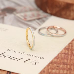 May include: A gold-colored ring with a white and iridescent inlay is the focal point of this image. Two other rings, one silver and one rose gold, are slightly out of focus in the background. The rings are on a cream-colored surface with the words "Merci beaucoup" and "bout Us" printed on it.