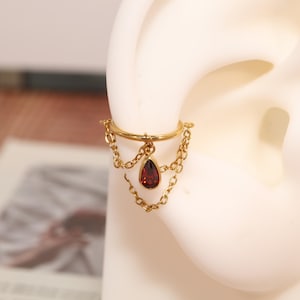 May include: A gold-toned ear cuff featuring a smooth circular band connected to two delicate chains that drape downwards. A single teardrop-shaped dark red gemstone charm hangs from the centre of the upper band, suspended by a small gold ring.