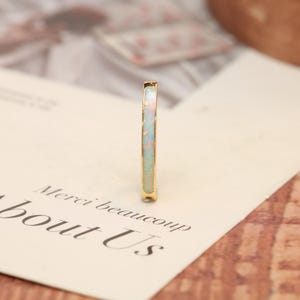 Könnte beinhalten: Ein goldener Ring mit einem Opal-Inlay. Der Opal hat eine Mischung aus Farben, darunter Weiß, Rosa und Grün. Der Ring ist vor dem Hintergrund eines weißen Papiers mit den Worten "Merci beaucoup about Us" platziert.