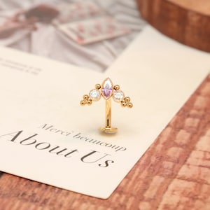 Könnte beinhalten: Ein goldfarbenes Labret-Piercing mit einem geschwungenen Zieraufsatz, besetzt mit einem zentralen, irisierenden Stein im Marquise-Schliff, zwei runden irisierenden Seitensteinen und kleinen goldenen Kugeln in Gruppen an den Außenrändern.