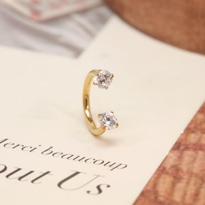 May include: A gold-colored, curved earring with two clear, round-cut gemstones. The earring is positioned vertically, with one gemstone at the top and one at the bottom. The background includes text that reads "Merci beaucoup Out Us".
