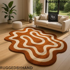May include: A brown and cream-coloured, wavy-patterned rug. The rug is placed on a wooden floor in a living room setting. The rug's design features a flowing, organic shape with a contrasting cream-coloured pattern. The words "RUGGEDBYHAND" are at the top and bottom of the image.