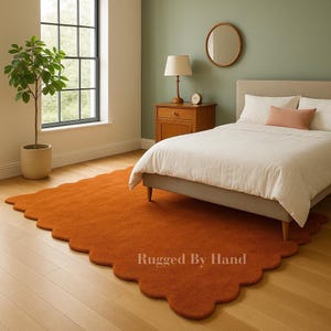 May include: An orange, scalloped-edge rug in a bedroom setting. The rug is placed under a bed with white bedding. A green plant and a window are visible in the background. The text "Rugged By Hand" is printed on the rug.