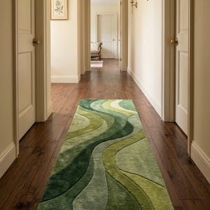 May include: A rectangular runner rug featuring a layered, wavy pattern in various shades of green, ranging from deep forest green to light sage and lime. The rug has a soft, textured pile and is positioned centrally on a dark wood floor.