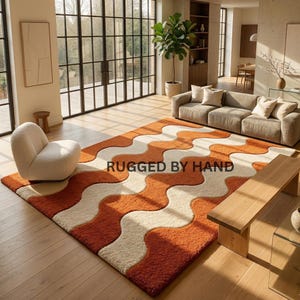Könnte beinhalten: Ein orangefarbener, cremefarbener und rostfarbener Teppich mit einem wellenförmigen Muster. Der Teppich trägt den weißen Schriftzug "RUGGED BY HAND". Der Raum umfasst ein Sofa, einen Stuhl und eine Holzbank, mit großen Fenstern im Hintergrund.