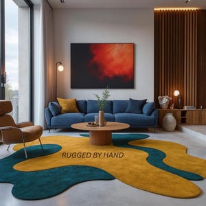May include: A modern living room featuring a blue sofa, a round wooden coffee table, and a large abstract painting. A unique rug in teal and mustard yellow with the text "RUGGED BY HAND" adds a pop of color and texture to the space.