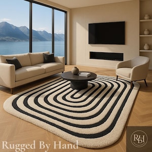 May include: A modern living room featuring a beige sofa, a black coffee table, and a cream-colored armchair. The focal point is a large, cream-colored rug with black, curved stripes. A large window offers a scenic view of mountains and water. The text "Rugged By Hand" is visible.