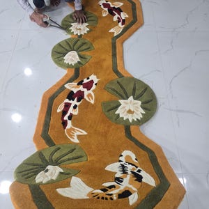 May include: An irregularly shaped rug featuring a mustard yellow background with a pattern of white koi fish with black and red markings, interspersed with green lily pads and white lotus flowers, finished with a dark green border.