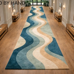 May include: A long, rectangular rug with a wavy pattern in shades of blue, beige, and cream. The rug is placed in a hallway with wooden floors and matching wooden tables. The text "RUGGED BY HAND" is at the top.