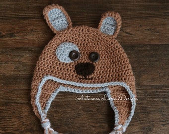 May include: A brown crocheted hat with a dog face design. The hat has two floppy ears, a brown nose, two black buttons for eyes, and a gray patch over one eye. The hat has a long braided tie with white and brown yarn.