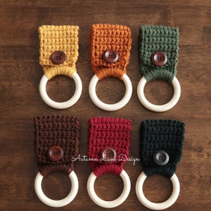 May include: Nine crocheted towel rings with wooden rings. The rings are in various colors, including yellow, orange, green, brown, red, black, white, and gray. Each ring has a wooden button in the center.