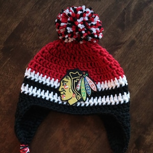 May include: A red, white, and black crocheted hat with a pom-pom on top and ear flaps. The hat features a black and red embroidered patch with a Native American headdress and the words "Blackhawks" on it.