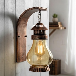 Może przedstawiać: Rustykalna lampa ścienna z miedzianym wykończeniem. Oprawa posiada zakrzywione ramię i wiszącą lampę w stylu latarni z przezroczystą szklaną żarówką. Widoczny jest tekst "AVhomedeco".