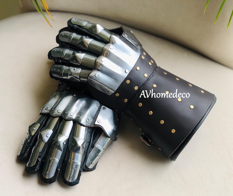 May include: A pair of medieval-style gauntlets. The gauntlets feature articulated metal plates for the fingers and knuckles, with a black leather cuff adorned with gold-colored rivets. The text "AVhomedeco" is visible on the cuff.