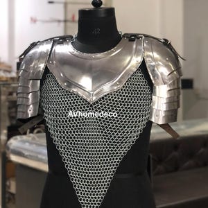 May include: A polished steel chest and shoulder armor set. The armor features layered shoulder plates, a central breastplate, and a chainmail lower section. The armor is displayed on a black mannequin. The text "AVhomedeco" is visible.