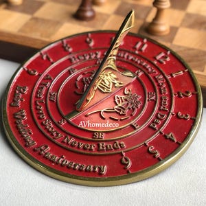 Pode incluir: Um relógio de sol vermelho e dourado com o texto "A True Love Story Never Ends" e "Happy Wedding Anniversary". O relógio de sol tem algarismos romanos e um gnomon dourado. O relógio de sol tem aproximadamente 15 cm de diâmetro.