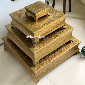 May include: A set of four gold-coloured, square decorative stands of varying sizes. Each stand features ornate floral and scrollwork patterns and rests on small, decorative feet. The stands are stacked, with the smallest on top.