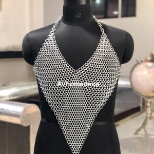 May include: A silver chainmail halter top displayed on a black mannequin. The top features a V-neck design and is constructed from small, interlocking metal rings. The mannequin is set against a neutral background.