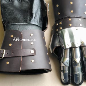 May include: A pair of medieval-style gauntlets. The gloves feature black leather palms and cuffs, with articulated metal finger and thumb protection. The cuffs are brown leather with brass rivets and a buckle closure. The text "AVhomedeco" is visible on the cuff.