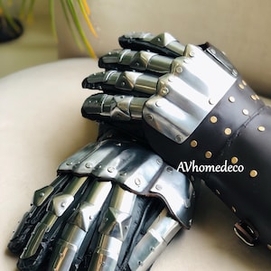 May include: Pair of medieval-style gauntlets. The gauntlets are made of polished metal plates and black leather. The metal plates are riveted together and have a shiny, reflective surface. The leather cuffs are adorned with gold-colored rivets. The text "AVhomedeco" is visible.