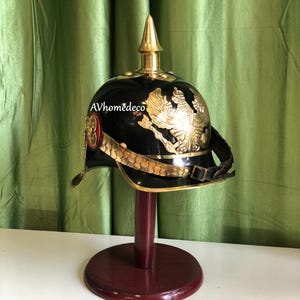 May include: A black and gold military helmet with a decorative eagle emblem and a gold spike on top. The helmet is displayed on a wooden stand. The text "AVhomedeco" is visible on the helmet.