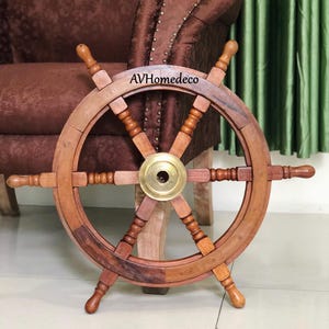 Op de afbeelding: Een houten scheepsstuurwiel met een messing middenstuk. Het wiel heeft meerdere spaken en een donkerbruine afwerking. De tekst "AVHomedeco" is zichtbaar op het wiel. Het wiel is een decoratief nautisch item.