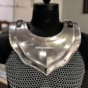 May include: A polished steel gorget, part of medieval armor, displayed on a black mannequin. The gorget has a curved shape to protect the neck and upper chest, with a chainmail lower section. The text "AVhomedeco" is visible.