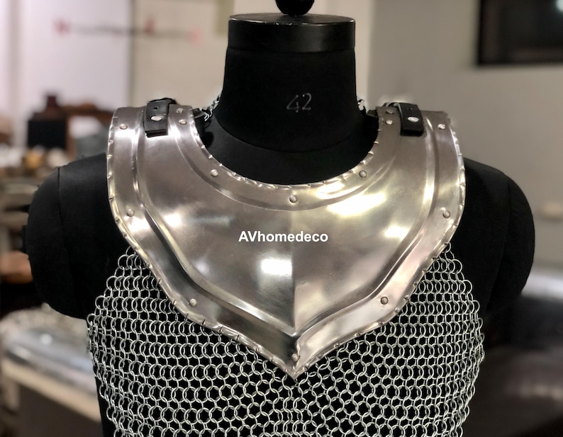 May include: A polished steel gorget, part of medieval armor, displayed on a black mannequin. The gorget has a curved shape to protect the neck and upper chest, with a chainmail lower section. The text "AVhomedeco" is visible.