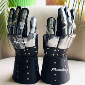 May include: Pair of medieval-style gauntlets. The gauntlets feature articulated metal finger plates and black leather cuffs adorned with brass rivets. The metal components are silver-toned, and the leather is black.