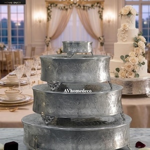 Può includere: Un set di alzatine per torte argentate con motivi floreali ornati. Il set comprende quattro alzatine rotonde di diverse dimensioni, ciascuna sostenuta da piedini decorativi. Sullo sfondo si vede una torta nuziale con decorazioni floreali.