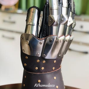 May include: A medieval-style gauntlet, featuring a combination of polished metal plates and dark brown leather. The metal plates are riveted to the leather, and the gauntlet has a flared cuff. The text "AVhomedeco" is visible.