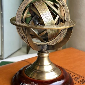 Puede incluir: Una esfera armilar de latón con símbolos del zodiaco y marcas de grados, montada sobre una base de madera. La esfera presenta un diseño detallado con los textos "TAURUS" y "GEMINI". La base es de color marrón oscuro con un vástago de latón.