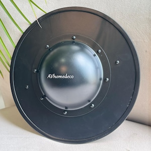 May include: A black metal shield with a rounded shape and a central, raised boss. The shield has a series of rivets around the boss and the outer edge. The text "AVhomedeco" is visible on the shield.