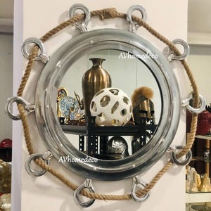 May include: A circular mirror with a silver frame and a nautical theme. It features a rope and metal ring border. The mirror reflects decorative items, including a vase and a white sphere. The text "AVhomedeco" is visible on the mirror's surface.