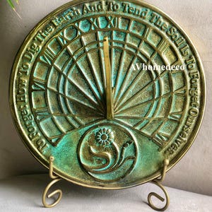 May include: A round, antique-style sundial with a verdigris finish, mounted on a decorative stand. The sundial features Roman numerals and the inscription "How To Dig The Earth And To Tend The Soil Is To Forget Ourselves."