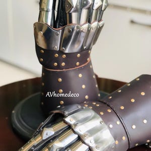 May include: Pair of medieval-style gauntlets. The gauntlets feature articulated metal plates over the fingers and knuckles, attached to a brown leather base. The leather is studded with small, gold-colored rivets.
