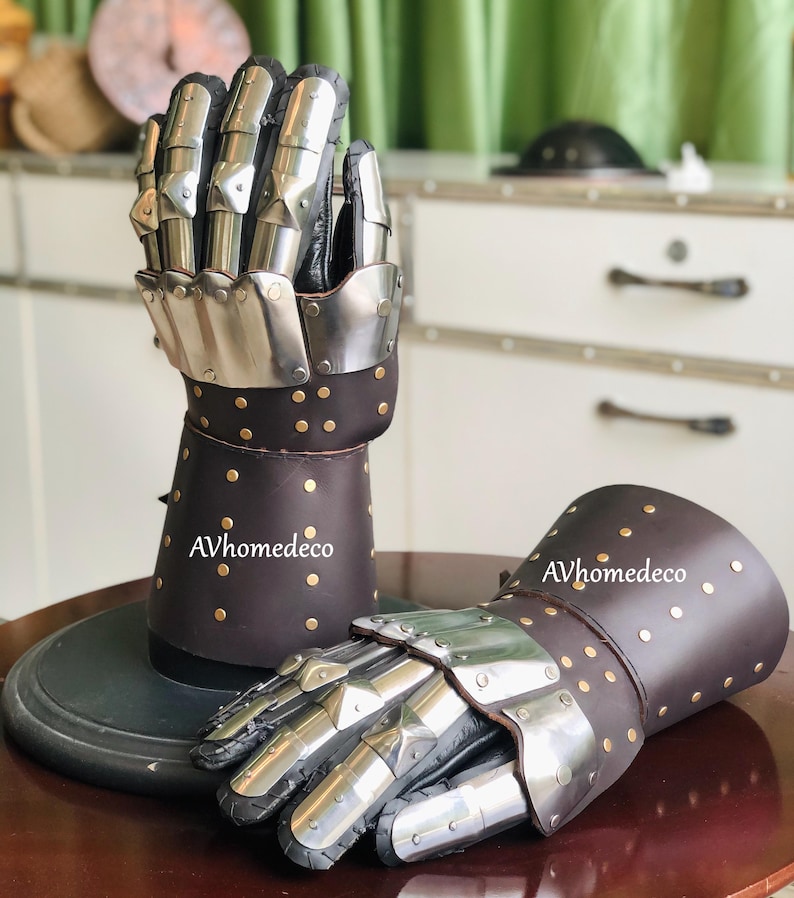 May include: A pair of medieval gauntlets. The gauntlets feature articulated metal finger plates and brown leather cuffs adorned with brass studs. The gauntlets are displayed on a dark surface. The text "AVhomedeco" is visible on the leather.