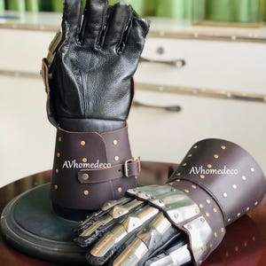 May include: A pair of medieval-style gauntlets. The gloves are black leather with brown leather cuffs, secured with buckles and rivets. The knuckles and fingers are covered with metal plates. The text "AVhomedeco" is visible on the cuffs.