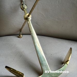 Puede incluir: Un ancla de latón con un diseño curvo, con un vástago largo y en ángulo y una aleta curva. El ancla tiene un acabado pulido y un pequeño anillo en la parte superior. El texto "AVhomedeco" es visible.