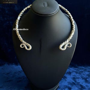 May include: A silver-colored, twisted metal collar necklace with a unique design. The necklace is displayed on a black mannequin head against a dark blue background. The ends of the necklace feature a decorative swirl.