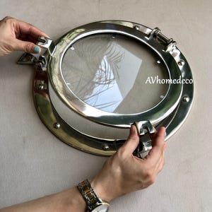 May include: A round, polished metal porthole with a clear glass window. The porthole is being opened by a person's hands. The porthole has a shiny, reflective surface and is mounted on a light-colored wall. The text "AVhomedeco" is visible.