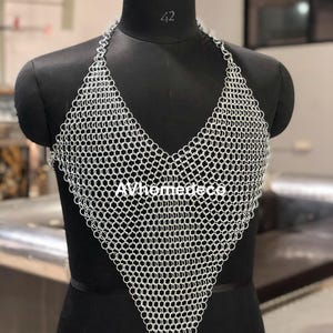May include: A silver chainmail halter top with a V-neck design. The top is displayed on a black mannequin. The text "AVhomedeco" is visible on the front of the top. The mannequin has a black base and a black head.