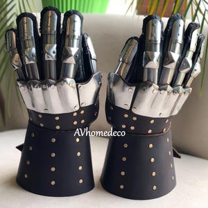 May include: Pair of medieval-style gauntlets. The gloves feature articulated metal finger plates and a black leather cuff with brass rivets. The gauntlets are designed to protect the hands and wrists.