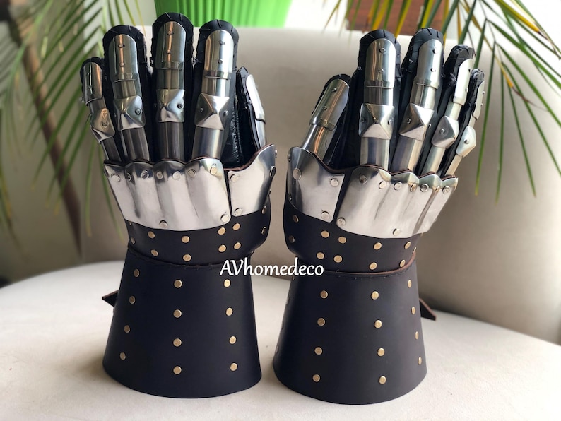 May include: Pair of medieval-style gauntlets. The gloves feature articulated metal finger plates and a black leather cuff with brass rivets. The gauntlets are designed to protect the hands and wrists.