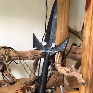 May include: A black metal spearhead with a central blade and two side blades. The spearhead is displayed on a wooden shelf with various necklaces and decorative items. The text "AVhomedeco" is visible.