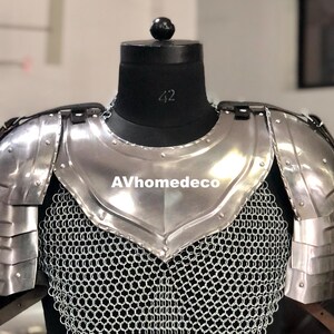 May include: A silver-colored metal chest and shoulder armor set, displayed on a black mannequin. The armor features layered shoulder plates and a chainmail shirt. The text "AVhomedeco" is visible on the chest plate.