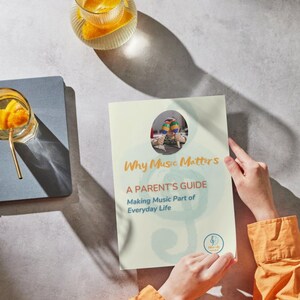 May include: A person holds a guide titled "Why Music Matters" with the subtitle "A Parent's Guide: Making Music Part of Everyday Life". The cover features a child with maracas. A glass of orange slices and a drink are in the background.