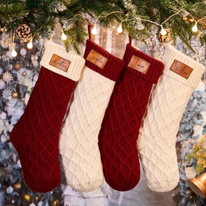 Peut inclure: Quatre bas de Noël personnalisés sont suspendus à une guirlande de pin et de lumières. Deux sont en tricot de câble bordeaux, et deux sont blancs avec un motif en losange. Chacun a une étiquette nominative en cuir.