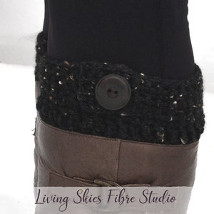 May include: Black crocheted leg warmers with a brown leather cuff and a large brown button. The leg warmers are made with a chunky yarn and have a textured look. The text "Living Skies Fibre Studio" is visible at the bottom of the image.